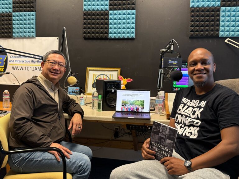 Two people in a studio smiling for camera, a laptop between them and one person holding a book.
