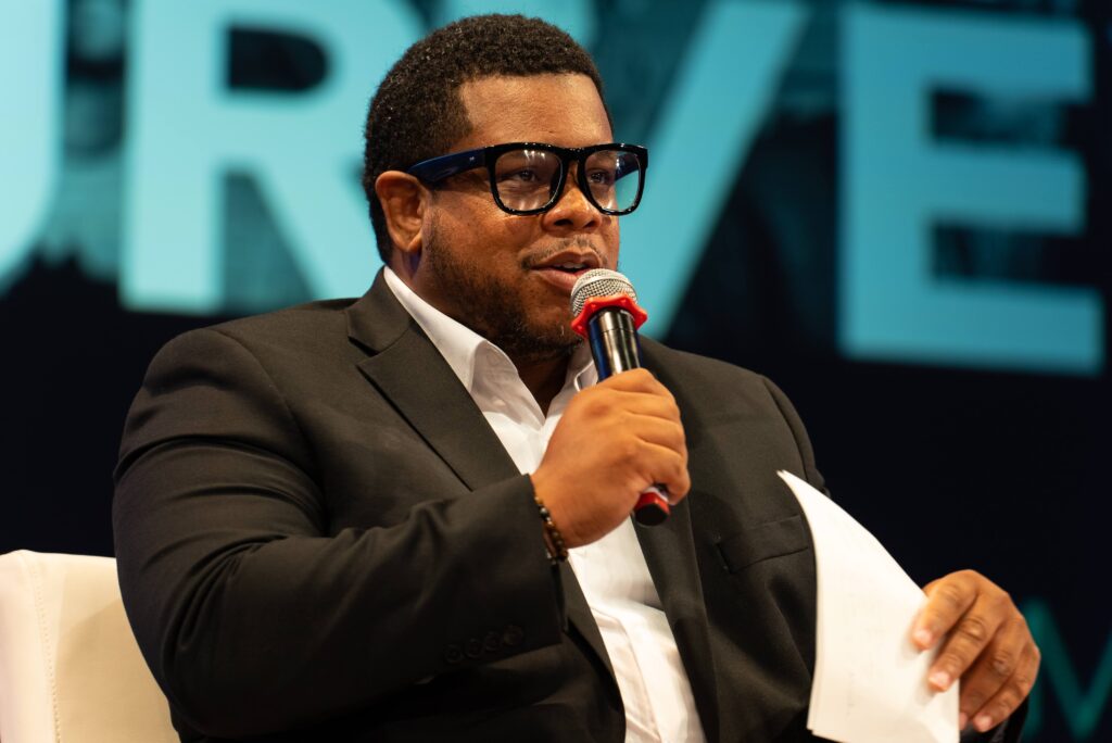 A man in glasses with a microphone speaks while holding a sheet of paper.