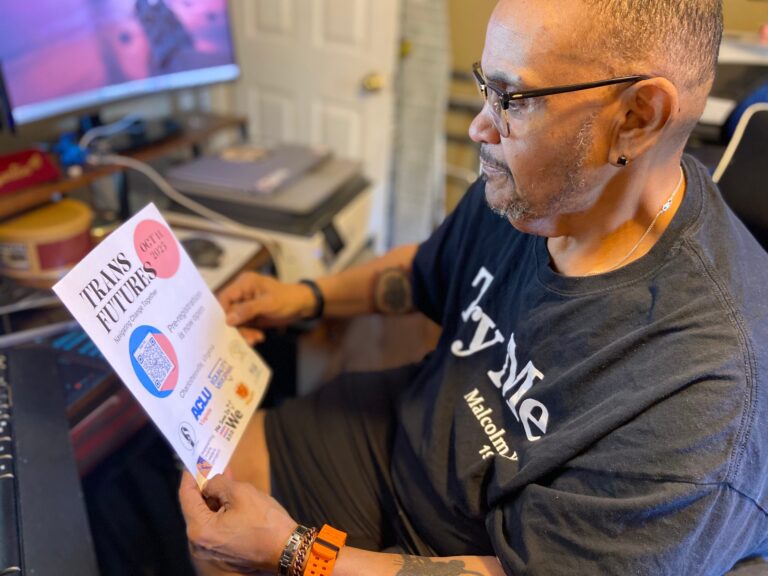 A man wearing glasses sits at desk in front of a computer looking at a flyer that reads "Trans Futures: Navigating change together."
