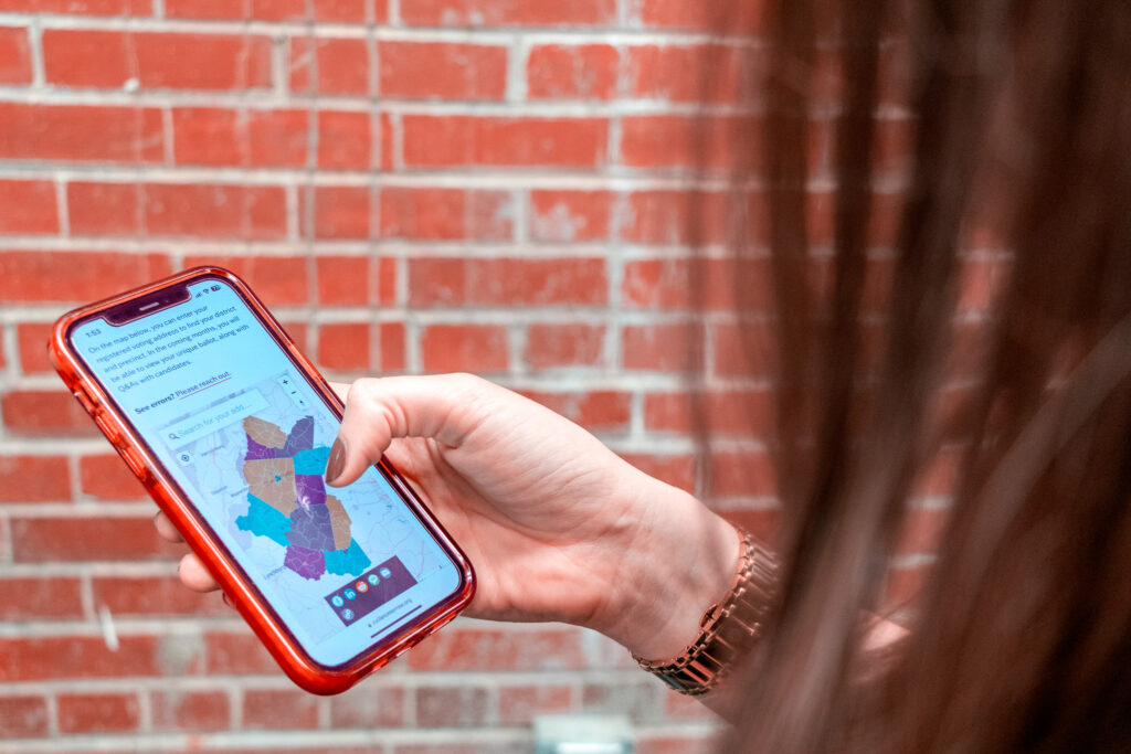 A hand with a watch on the wrist looking at a phone with a colorful map of central Virginia counties, brick wall in the background.