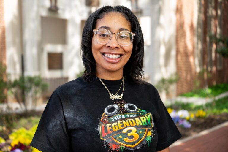 Woman in glasses and t-shirt standing in front of brick building and flowers smiles for camera. Her shirt has a colorful graphic that says "Free the legendary 3."