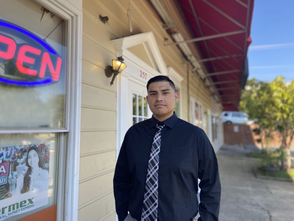 A man in a black shirt and tie stands in front of a store with part of a neon "Open" sign in the foreground.