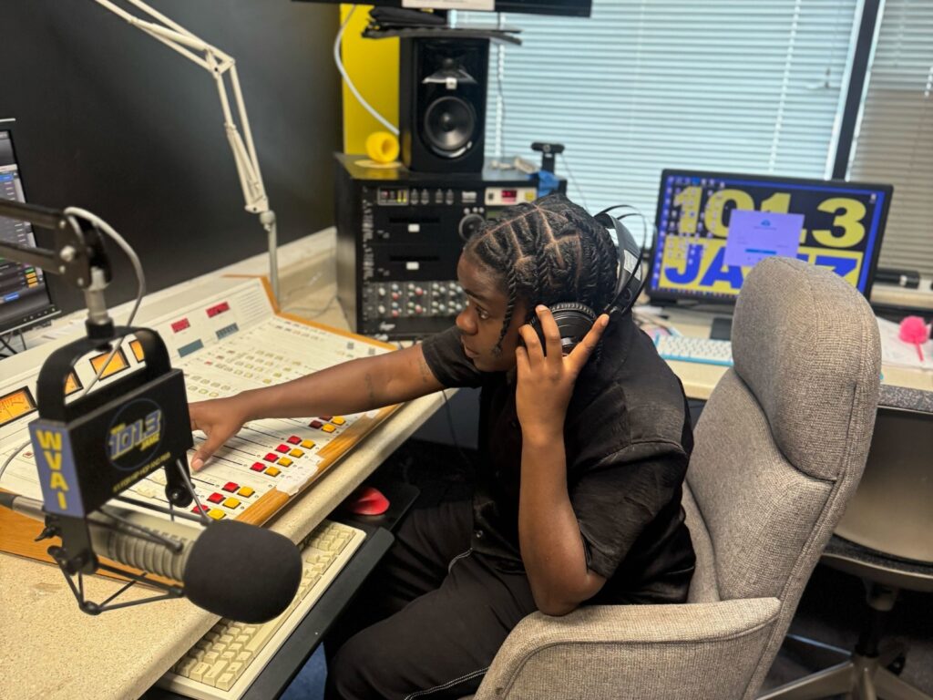 A person dressed in a black t-shirt and black jeans adjusts a fader on a sound mixing board. In the foreground is a suspended microphone that reads, "WVAI," and "101.3."