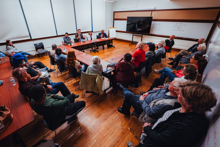 Two dozen people sit around conference tables in a classroom with hardwood flooring and whiteboards on the walls.